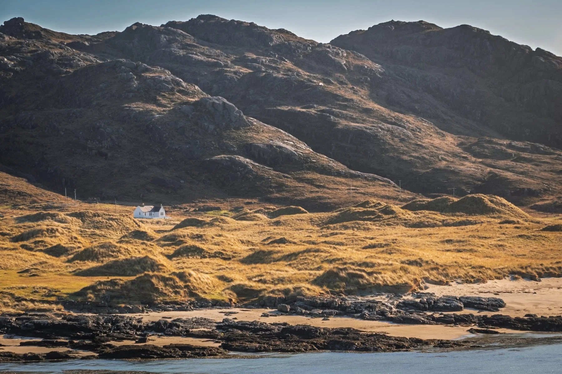 A croft house amongst sand dunes sitting beneath the rocky slopes of Meall Sanna, side lit by soft evening light | Ardnamurchan Scotland | Steven Marshall Photography