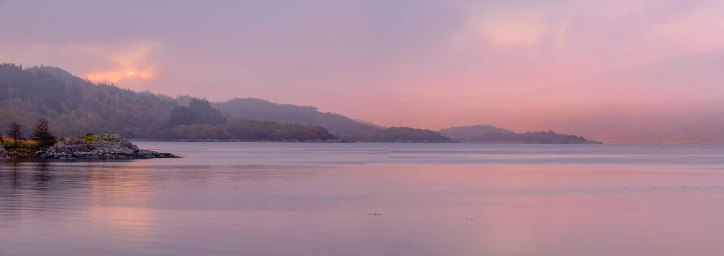 A peaceful Loch Sunart reflecting a pastel-coloured sky at dawn, with distant hills and trees lining the shoreline | Steven Marshall Photography
