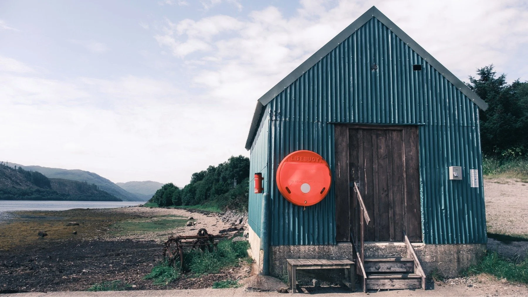 A green walled corrugated tin/iron building beside the sea with a red lifebuoy and wooden door on its gable end | Loch Sunart Strontian Ardnamurchan Scotland | Steven Marshall Photography
