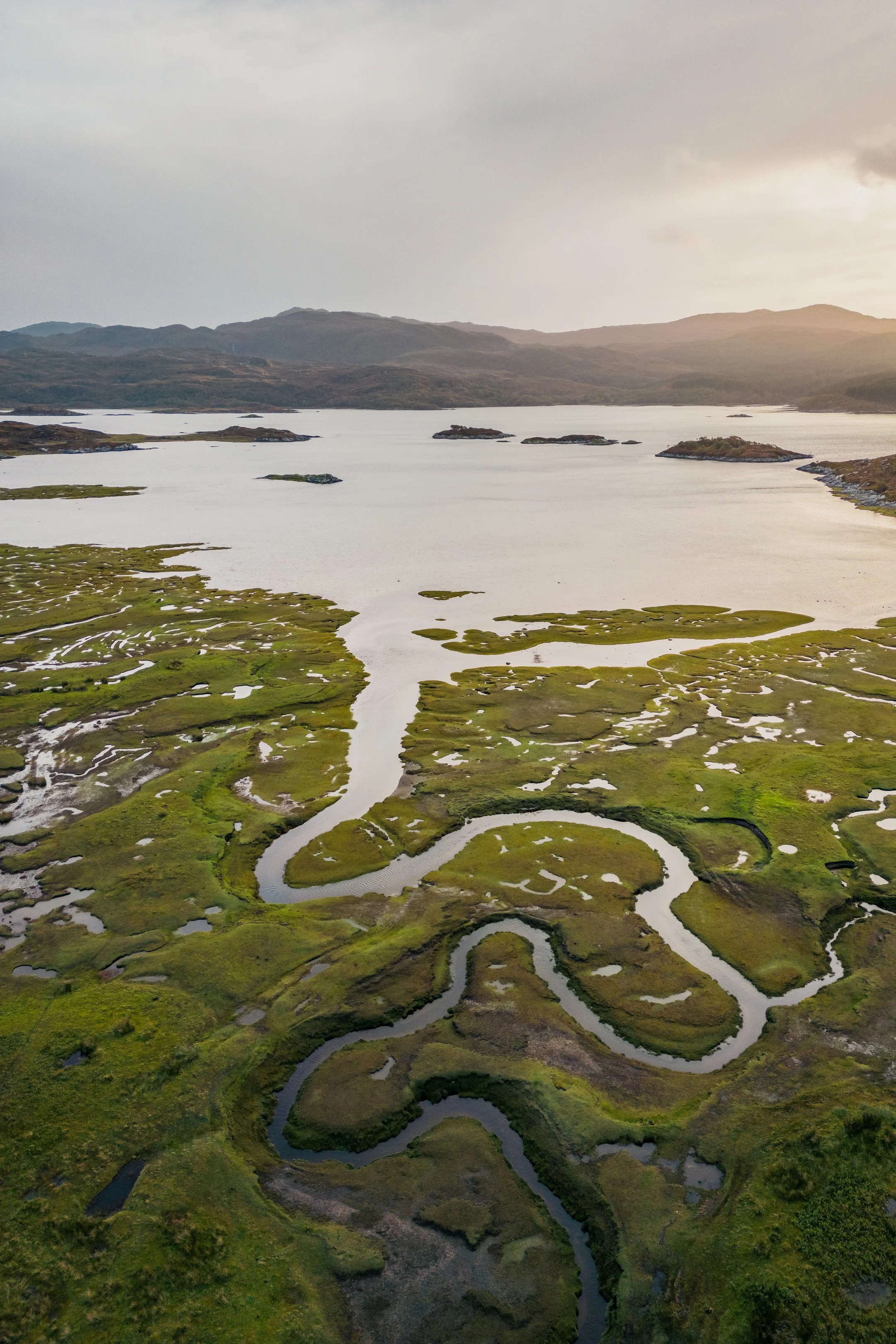 Aerial view of a winding Carnoch River meandering through the salt marsh at the head of Loch Sunart, with mountains in the background and a cloudy sky overhead | Steven Marshall Photography