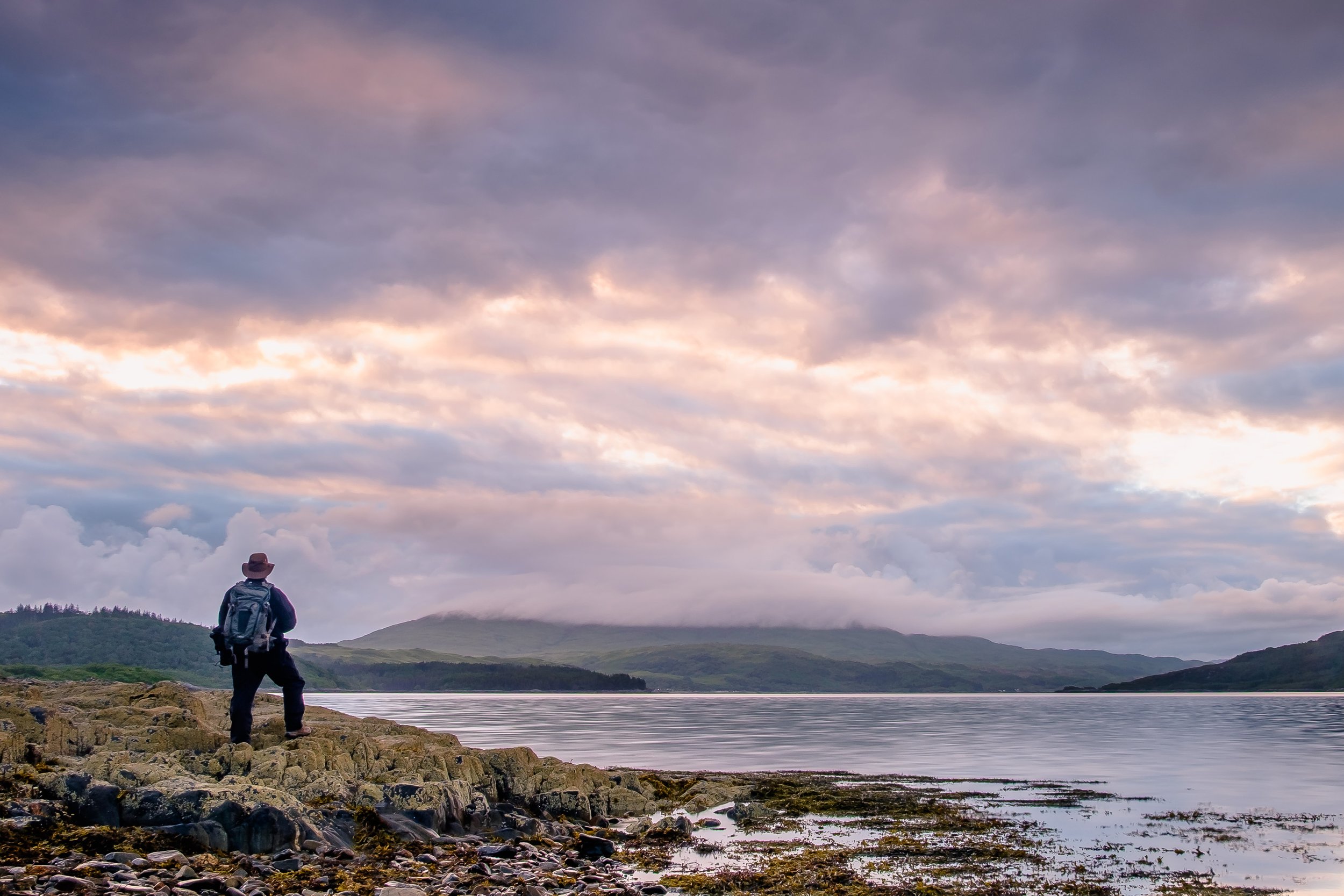 A person with a hat and backpack stands on rocky shoreline looking at a lake with mountains in the distance under a sky with pink and purple clouds during sunset or sunrise | Steven Marshall Photography