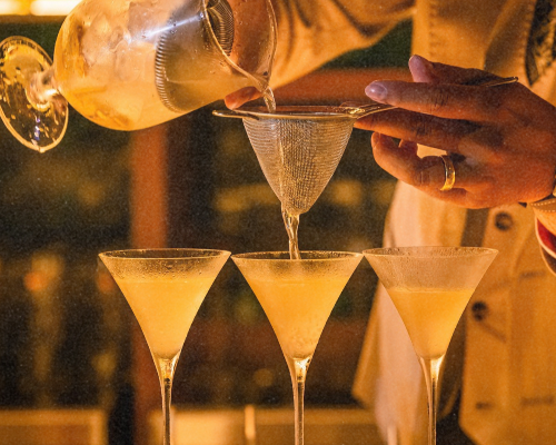 A woman with dark hair, gold hoop earrings, wearing a dark top, pouring a clear liquid from a bottle into a container, in a dimly lit bar or restaurant.
