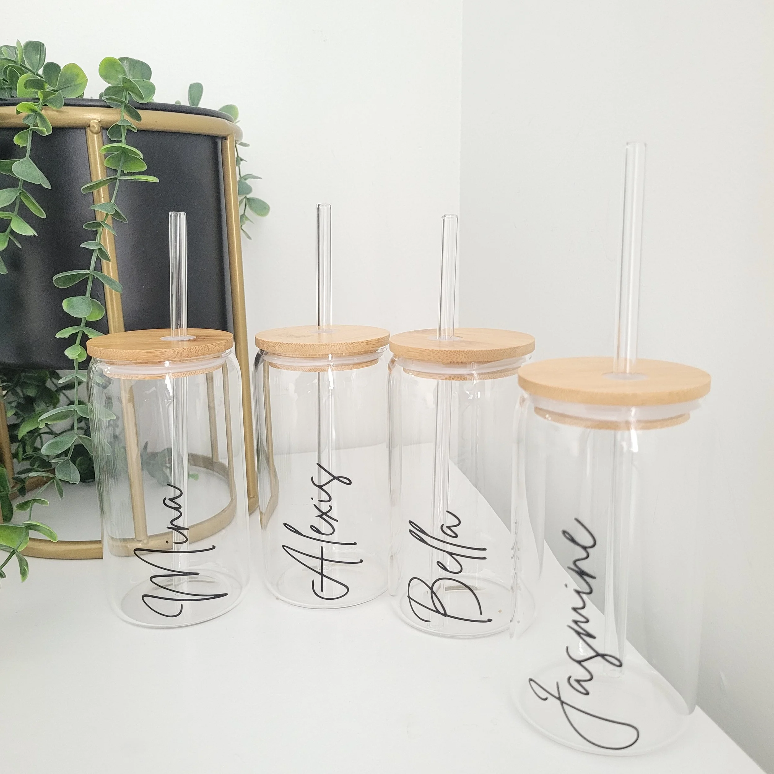Clear glass jars with wooden lids and black cursive writing on the surface, placed in a row against a white wall with another wall in the background, next to a green leafy plant.