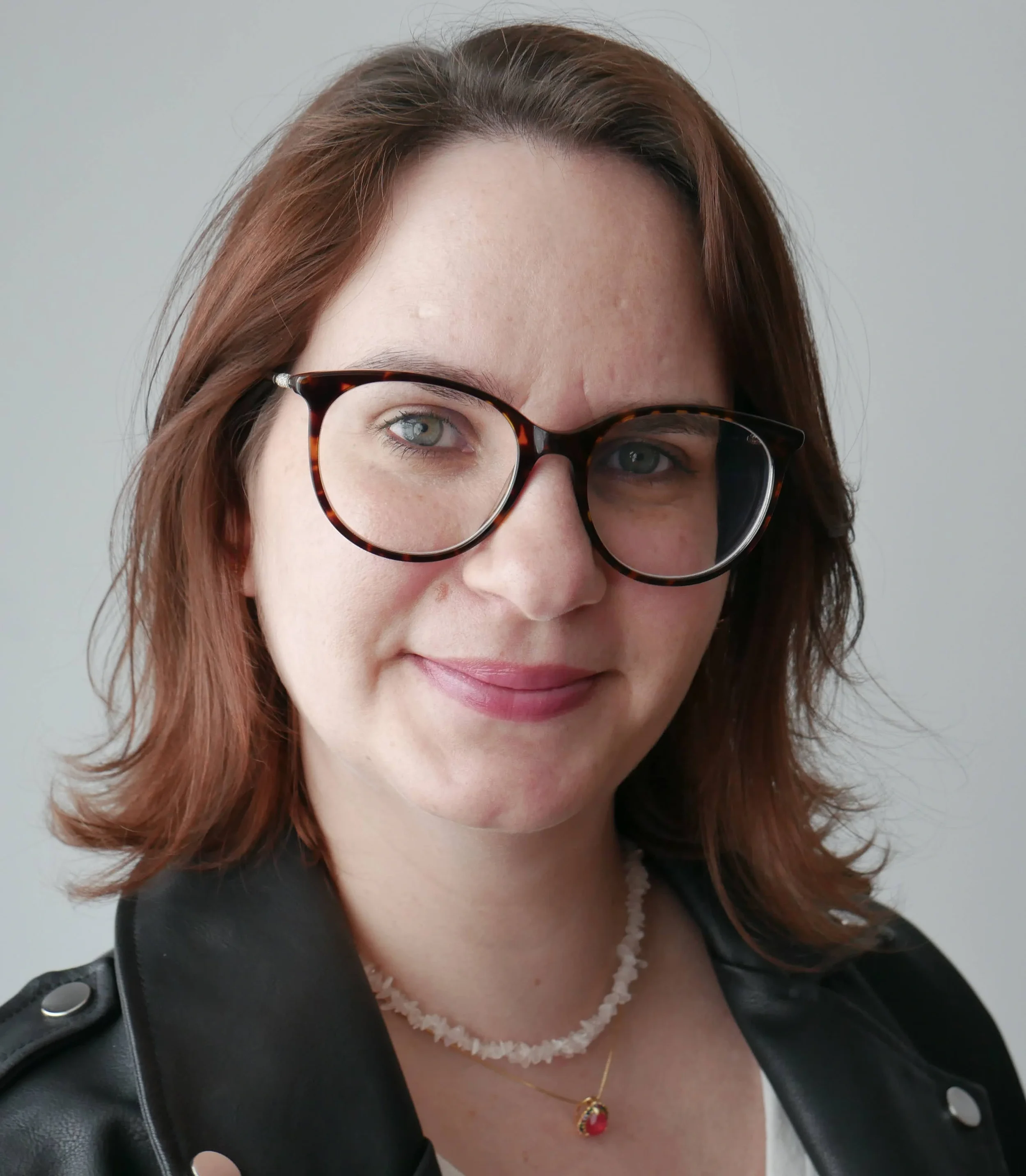 Portrait d'une femme avec des cheveux bruns, portant des lunettes à monture tortoise, un collier en perles blanches, une chaîne dorée avec pendentif rouge, et une veste en cuir noire, souriant doucement.