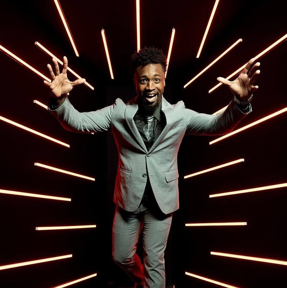 A man in a light gray suit with a black shirt and tie, smiling and reaching out with both hands, stands in front of a black background with red neon light strips radiating outward.