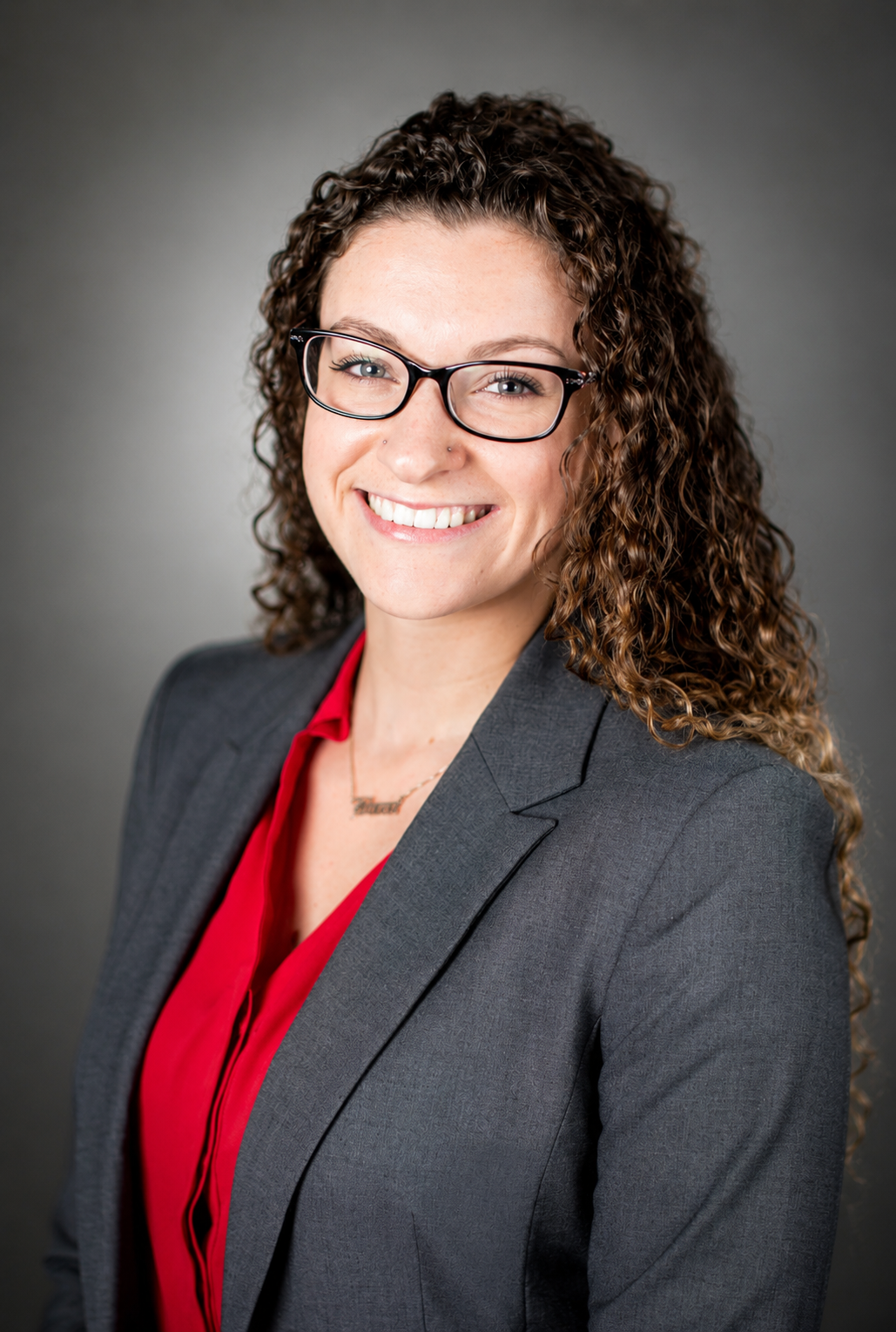 A woman with curly brown hair and glasses smiling, wearing a gray blazer over a red blouse, against a gray background.