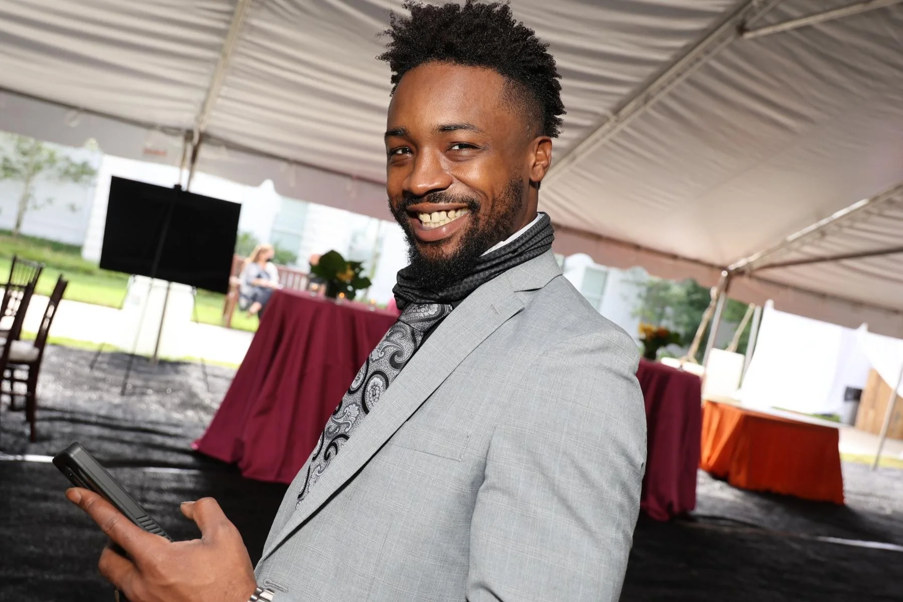 A man dressed in a light gray suit with a black and white patterned tie, smiling and holding a phone, at an outdoor event under a tent with tables and chairs in the background.