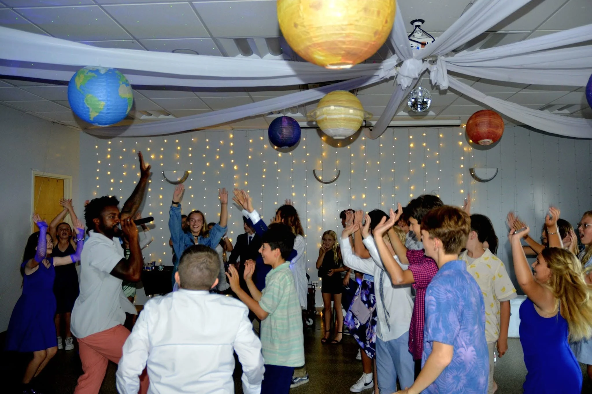 Group of people dancing and celebrating at a party with decorative planet and star globes hanging from the ceiling, white drapes, string lights, and a disco ball, in an indoor venue.