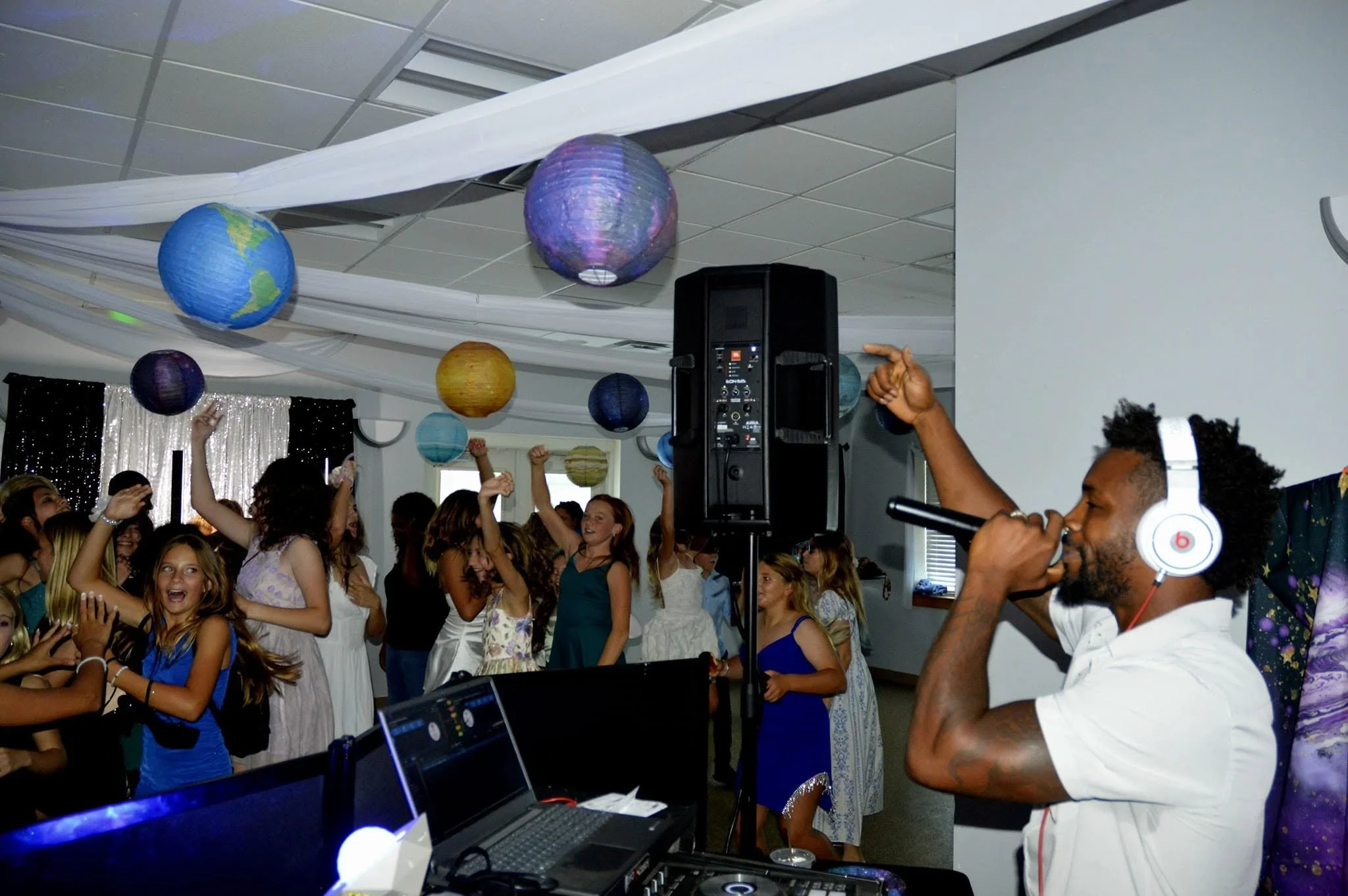 A lively indoor party with young people dancing and celebrating. Decor includes colorful paper lanterns resembling planets hanging from the ceiling. There is a DJ or music setup with a laptop and speakers, and a person wearing headphones appears to be DJing or managing music. The atmosphere is energetic and festive.