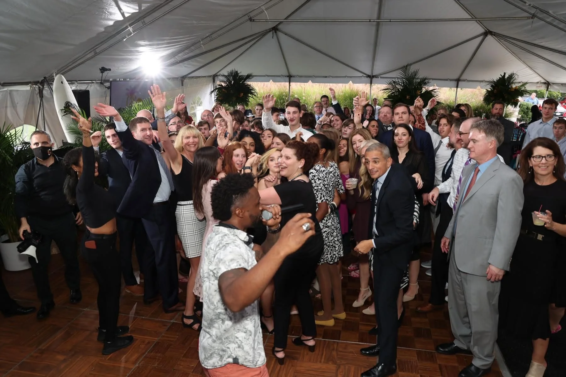 A large group of people dancing and socializing inside a decorated tent at a celebration or party. Many are smiling, with some waving and holding drinks. The setting appears festive with plants and a surfboard decoration visible.