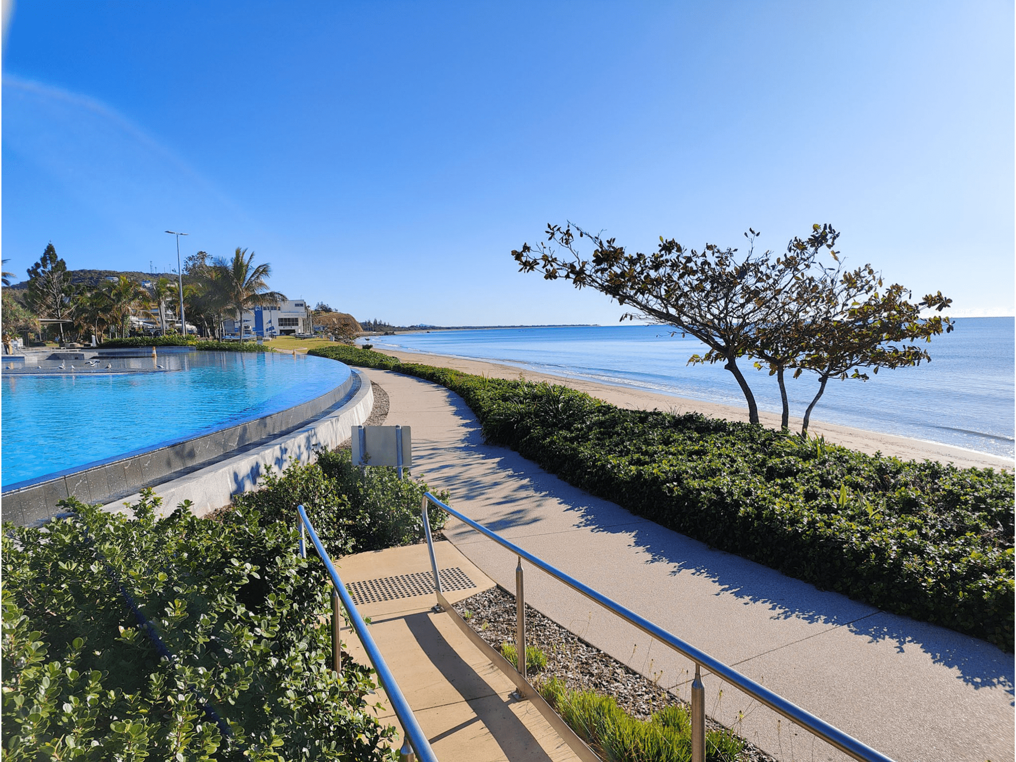 A scenic view of a coastal area with a swimming pool in the foreground, a sandy beach, and the ocean in the background. There are trees, shrubs, and modern buildings along the shoreline, under a clear blue sky.