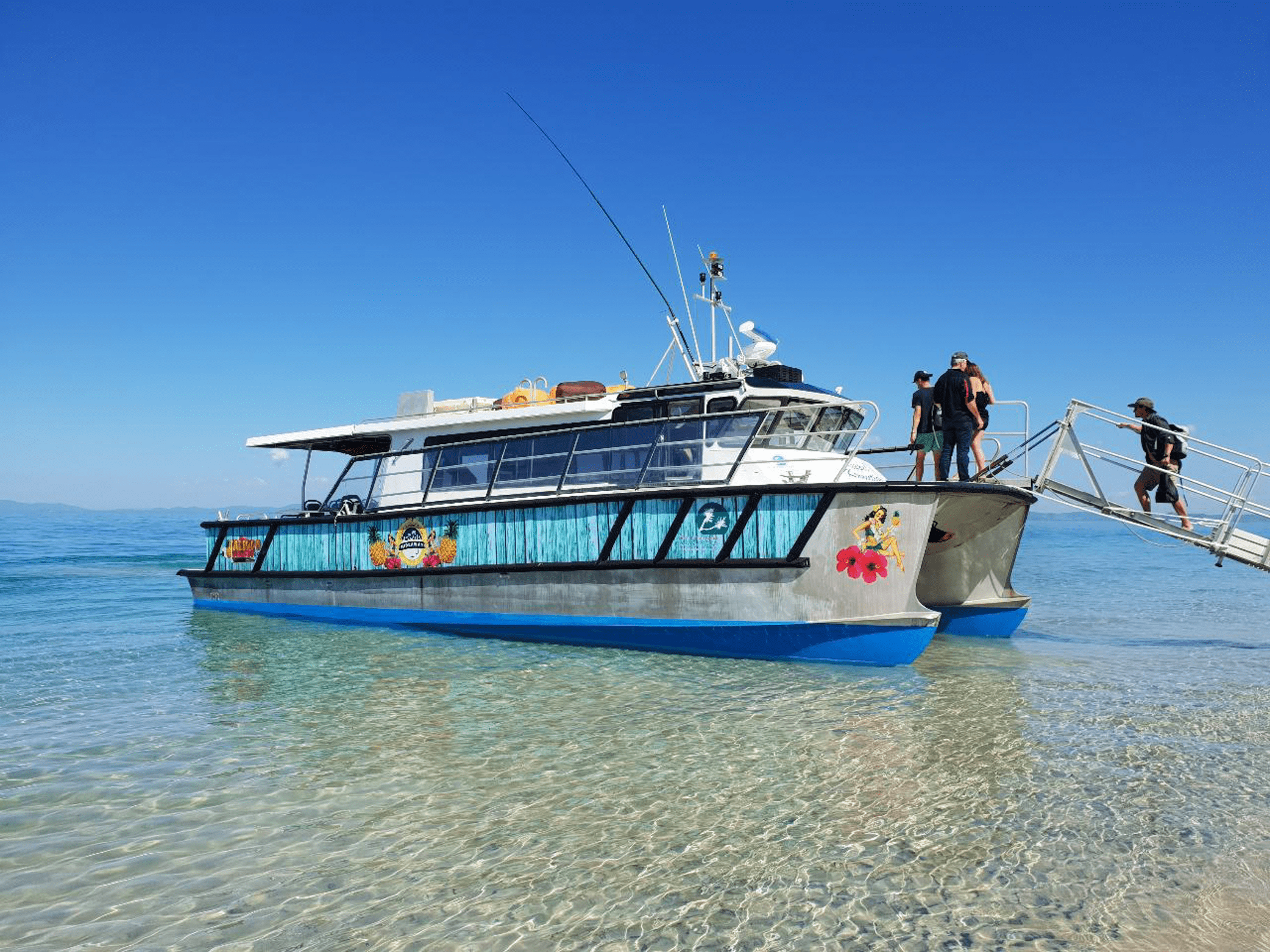 A boat with tropical decorations on its side floating on clear water, with people on the deck and a bright blue sky overhead.