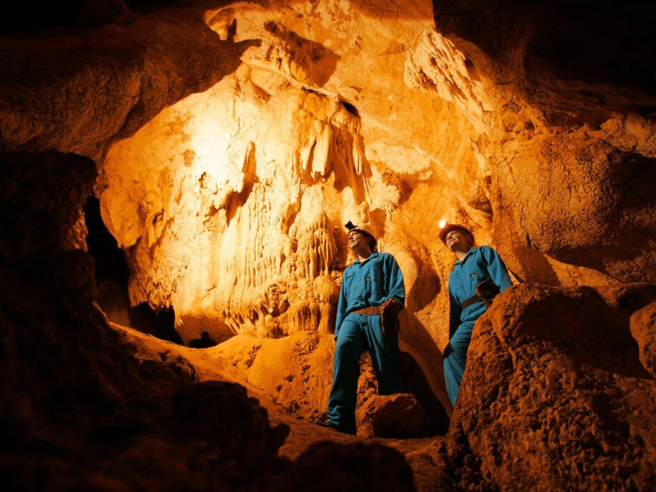 Two people in blue clothing and helmets exploring a large underground cave with stalactites and stalagmites illuminated by orange lighting.