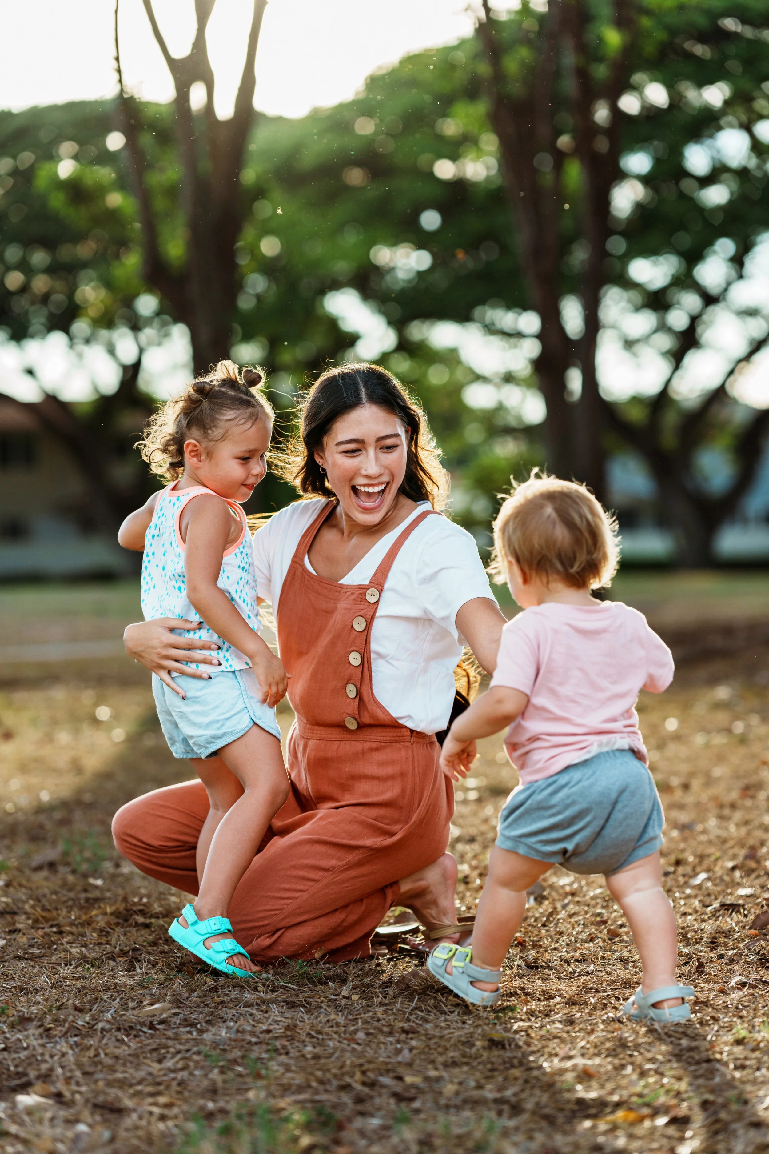 A woman with shoulder-length dark hair, wearing a white shirt with short sleeves and orange overalls, is kneeling on the ground outdoors, smiling and playing with two young children. One child, a girl with curly hair in pigtails, wearing a white and blue polka dot dress and bright blue sandals, is being held up by the woman. The other child, a boy with short light hair, wearing a pink shirt, gray shorts, and light blue sandals, is standing with his back to the camera. They are in a park with trees and sunlight filtering through the leaves.