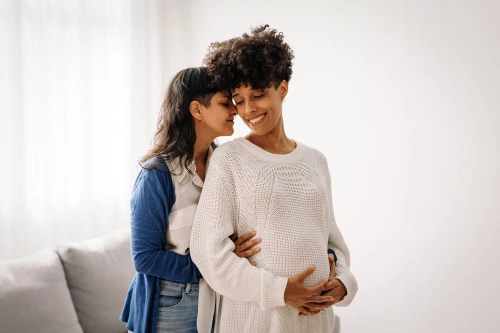 Same-sex couple standing face-to-face lovingly holding a pregnant belly, representing inclusive maternal mental health support across diverse family structures