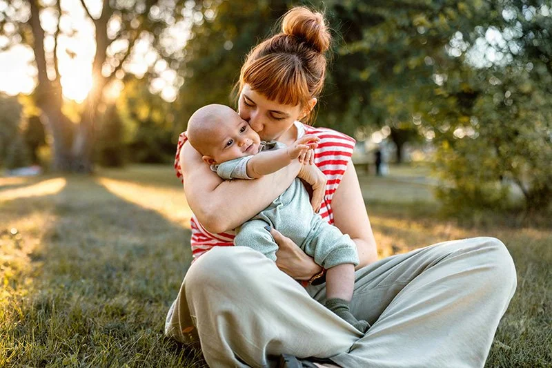 Mother gently kissing her baby in a park outdoors, representing postpartum therapy and early motherhood mental health support