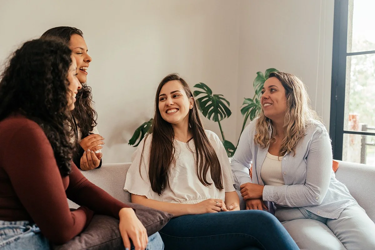Four women laughing and talking together on a couch, representing the supportive community and team of Bloomera perinatal psychologists