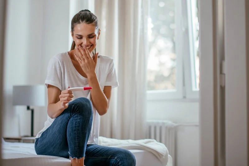 Smiling woman sitting on a bed reacting joyfully to a positive pregnancy test, representing preconception and fertility psychology support