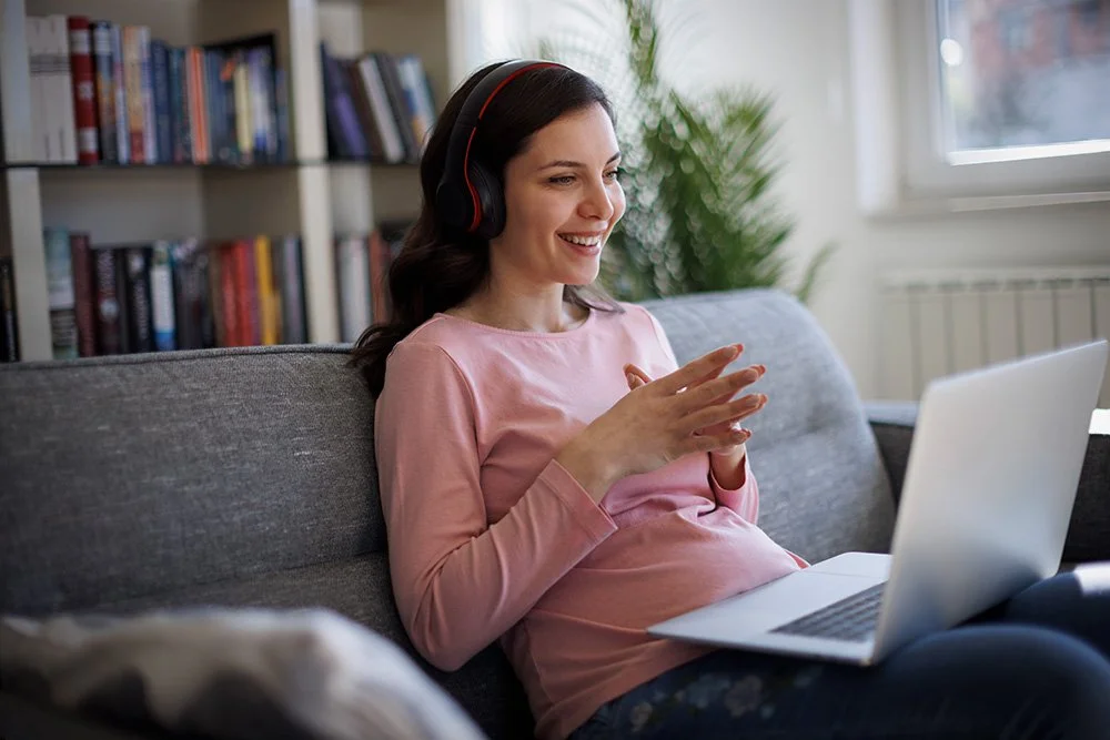 Pregnant woman wearing headphones smiling during an online telehealth psychology consultation on her laptop