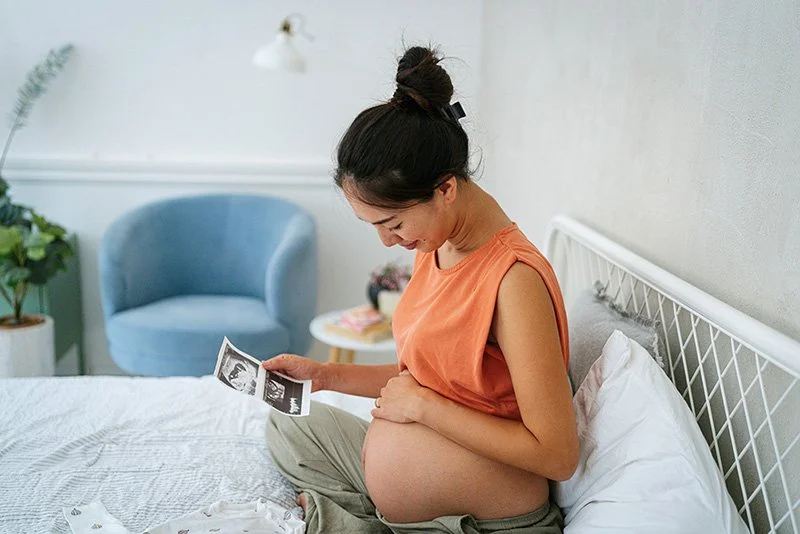 Pregnant woman sitting on a bed looking at ultrasound photos, representing pregnancy psychology and prenatal anxiety support