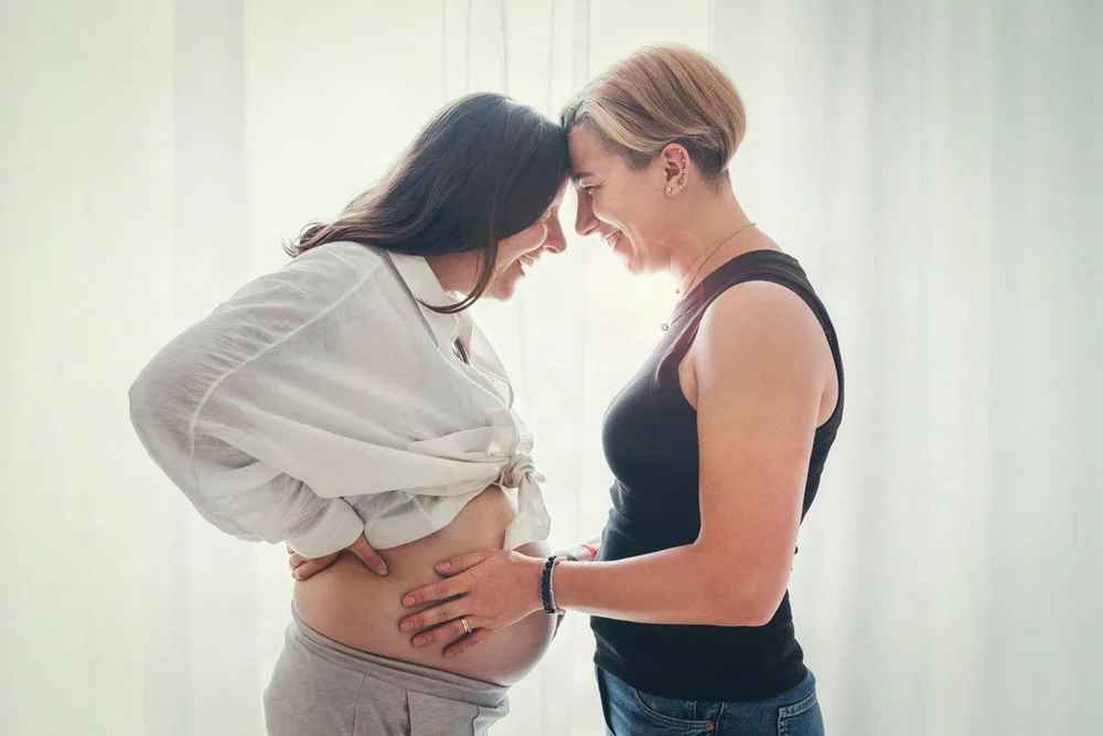 wo women standing face to face holding a pregnant belly, representing maternal support and perinatal care across identities, sexualities and family structures