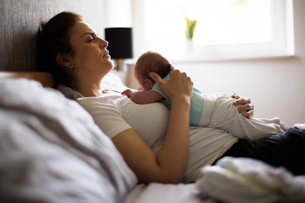 Exhausted mother lying on a bed with her newborn resting on her chest, representing postpartum therapy and early bond support