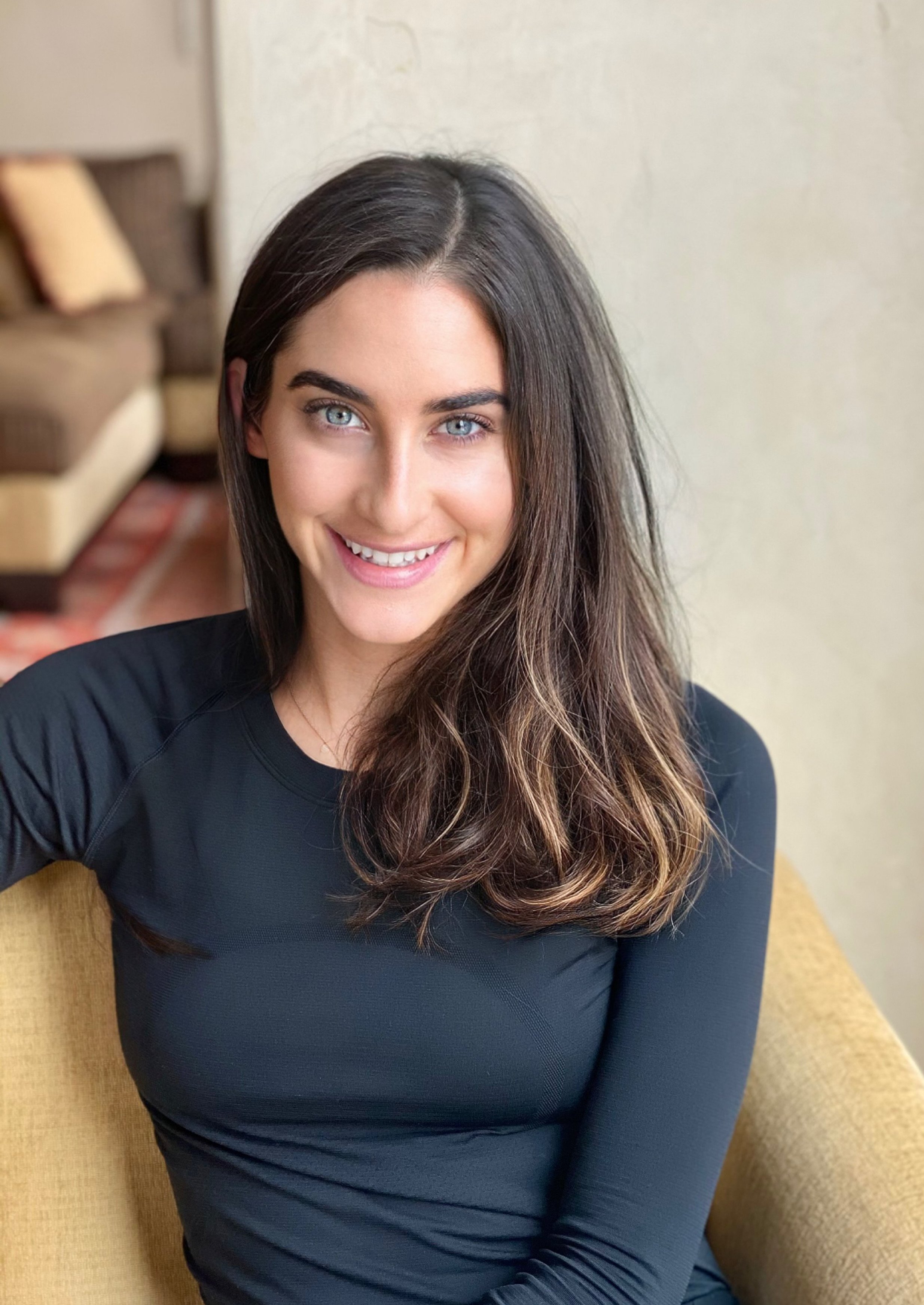 A woman with long, wavy brown hair and blue eyes sitting on a yellow couch, smiling at the camera.