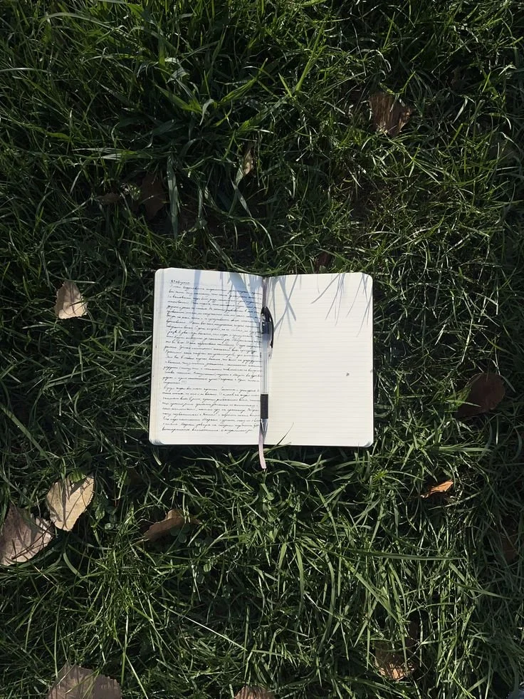 Open notebook with handwriting and a black pen placed on the grass with fallen leaves.