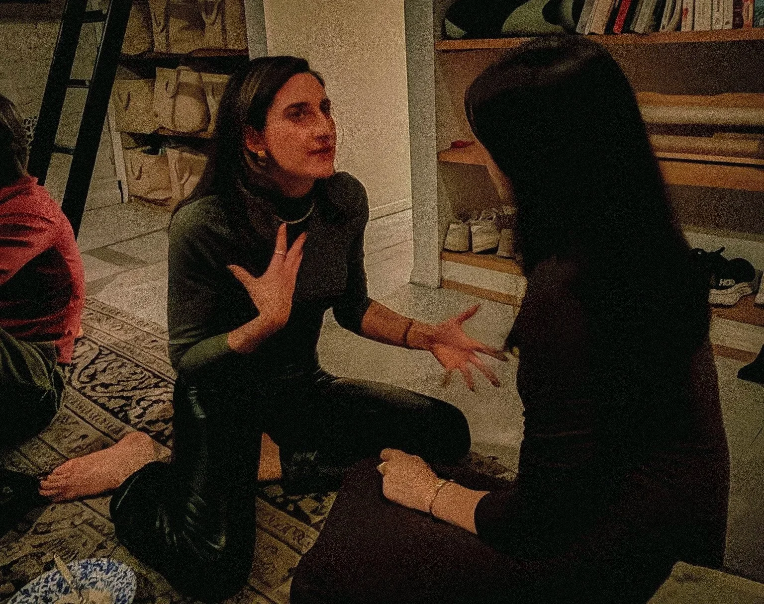 Two women having a conversation indoors. One woman is sitting on the floor, gesturing with her hands, while the other woman is sitting on a chair, listening. The background includes bookshelves and a patterned rug.