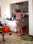 A woman is cooking in a kitchen while standing by the stove, with a open cupboard above. There is a chair with a red pillow nearby and a vacuum cleaner on the floor.