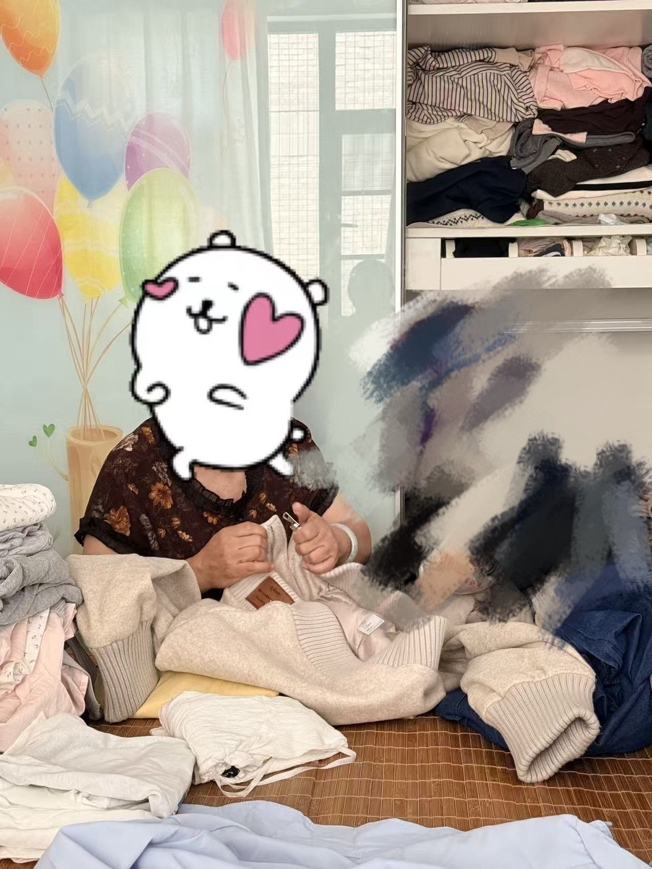 A person sitting on a bed with piles of clothes around, holding a piece of clothing, with their face covered by a cartoon bear sticker. Behind them is a wall with colorful balloons and a window with a grid pattern.