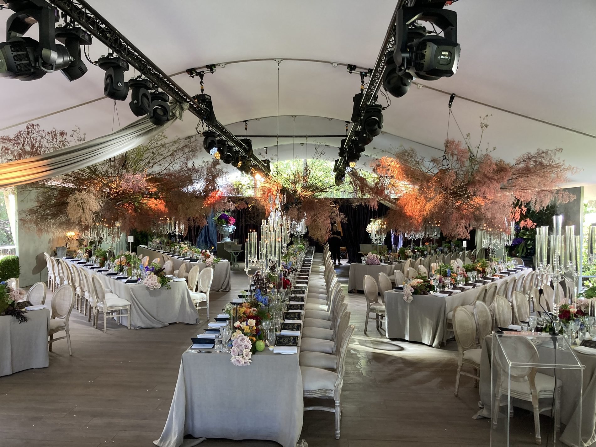 Elegant banquet hall decorated with peach-colored floral arrangements overhead, long tables set with floral centerpieces, candles, and place settings, surrounded by white chairs, with lighting and drapery accents.