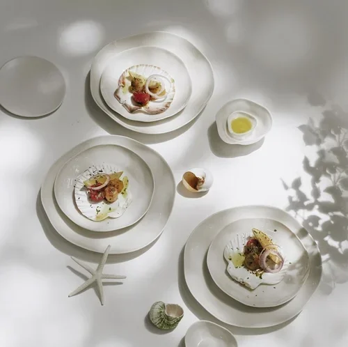 Seafood dishes on white plates with lemon wedges, starfish, and seashell decor on a white table with shadows from nearby foliage.