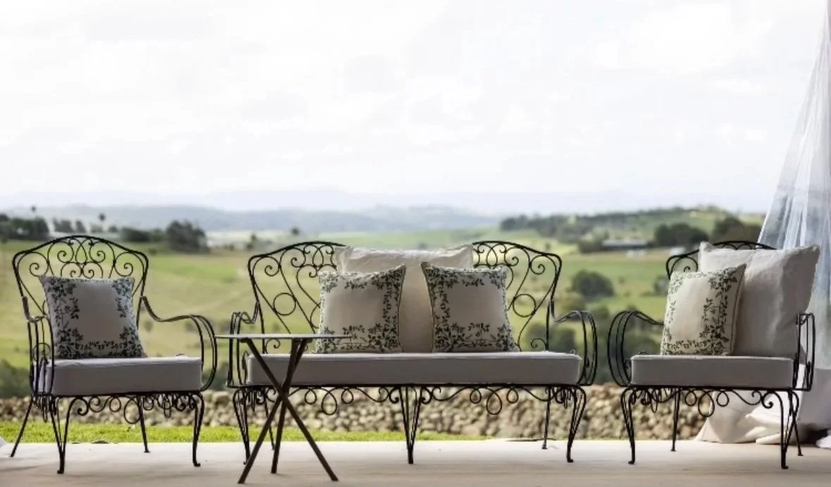 A porch with black iron patio furniture, including a bench and two chairs with cushions, overlooking a scenic countryside with rolling hills and fields.