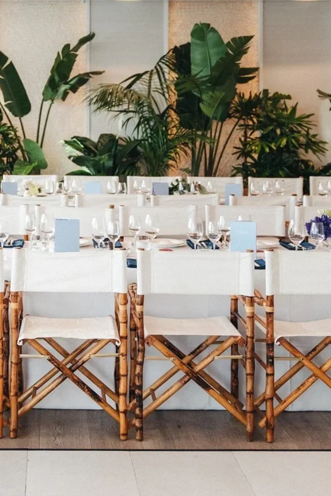 A dining setup with tables, white chairs, and green tropical plants in the background, indicating a decor for a special event or restaurant.