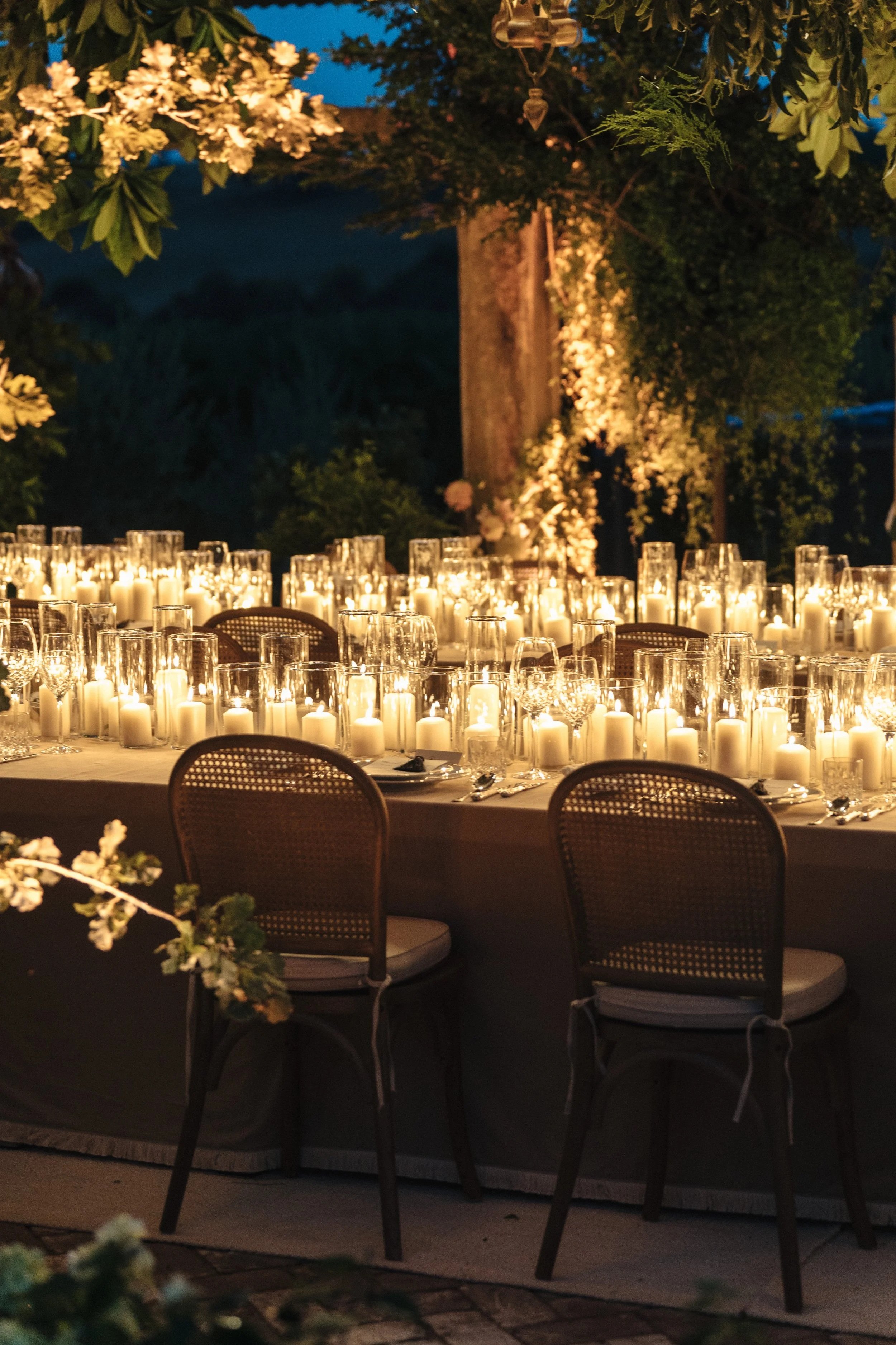 A nighttime outdoor dining setting with a long table decorated with numerous lit white candles in glass holders. Behind the table, there is a backdrop of lush greenery and flowers, with lighting illuminating the scene and the dark blue evening sky vi