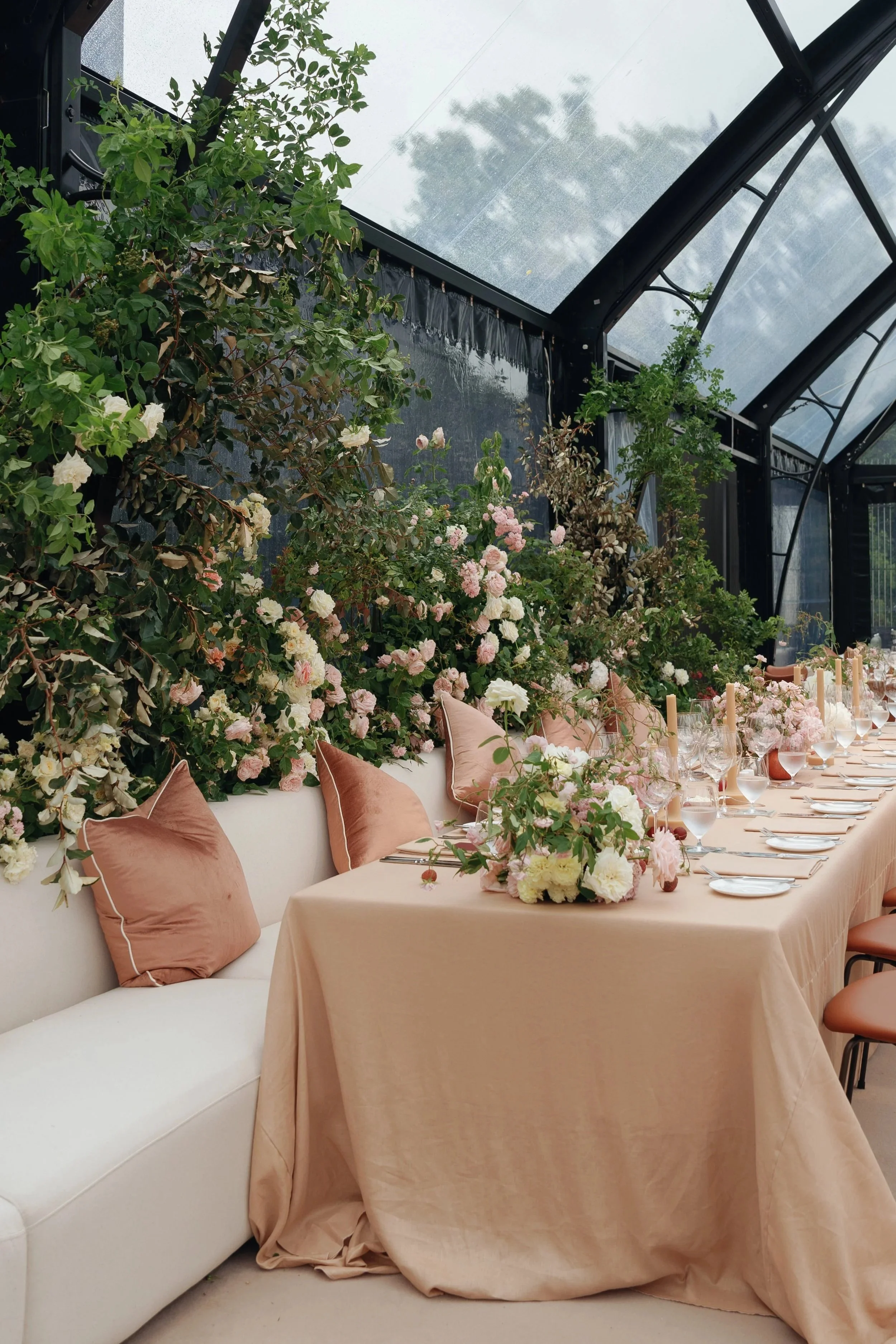 Elegant dining table set with pink tablecloth, surrounded by pink velvet pillows on a white sofa, adorned with floral arrangements of pink and white flowers, set in a bright conservatory with large glass windows and lush green foliage outside.