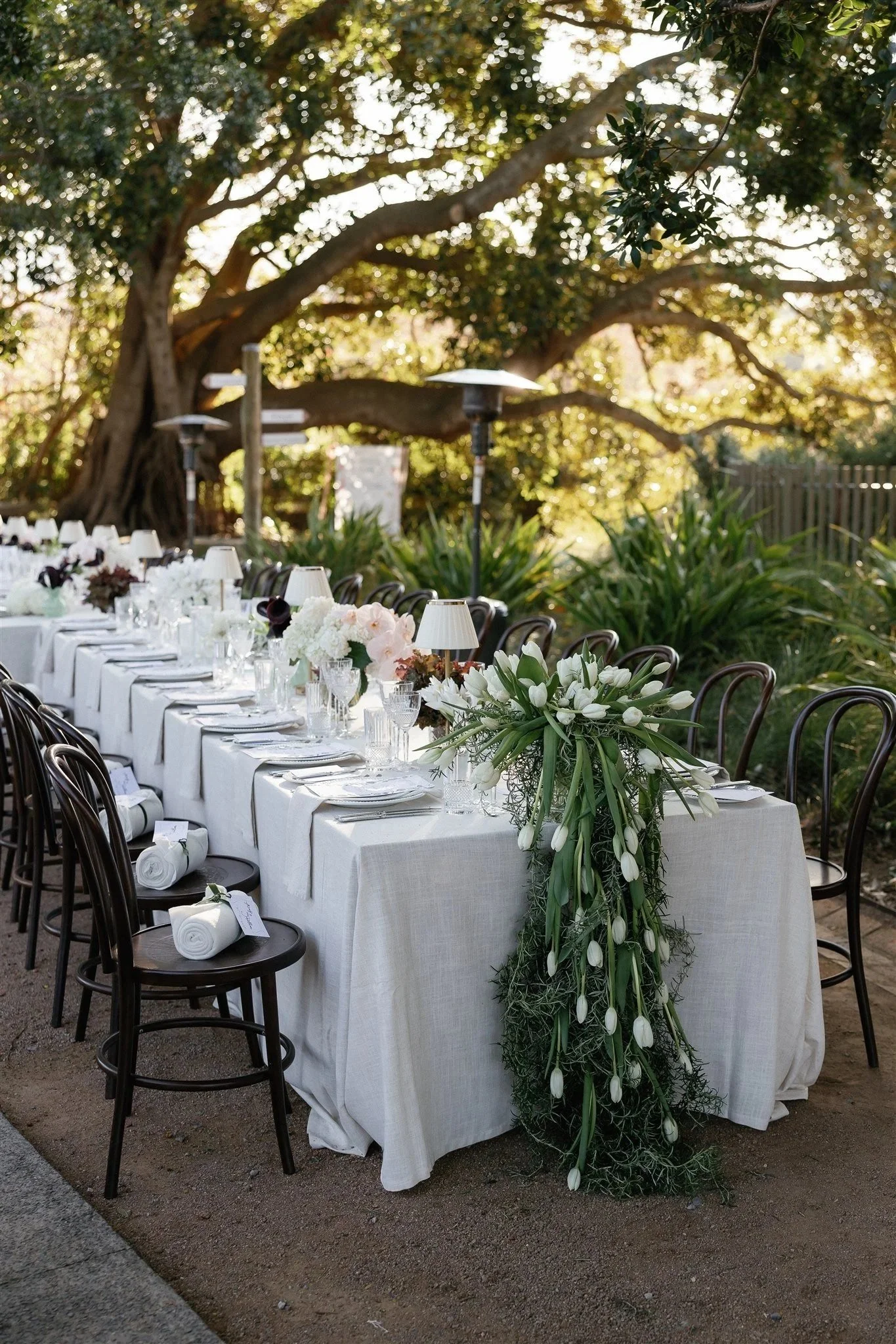 Long banquet table with white tablecloths decorated with floral centerpieces and white rolled napkins, set outdoors under trees with hanging lights, black chairs around the table, and a wooden fence in the background.