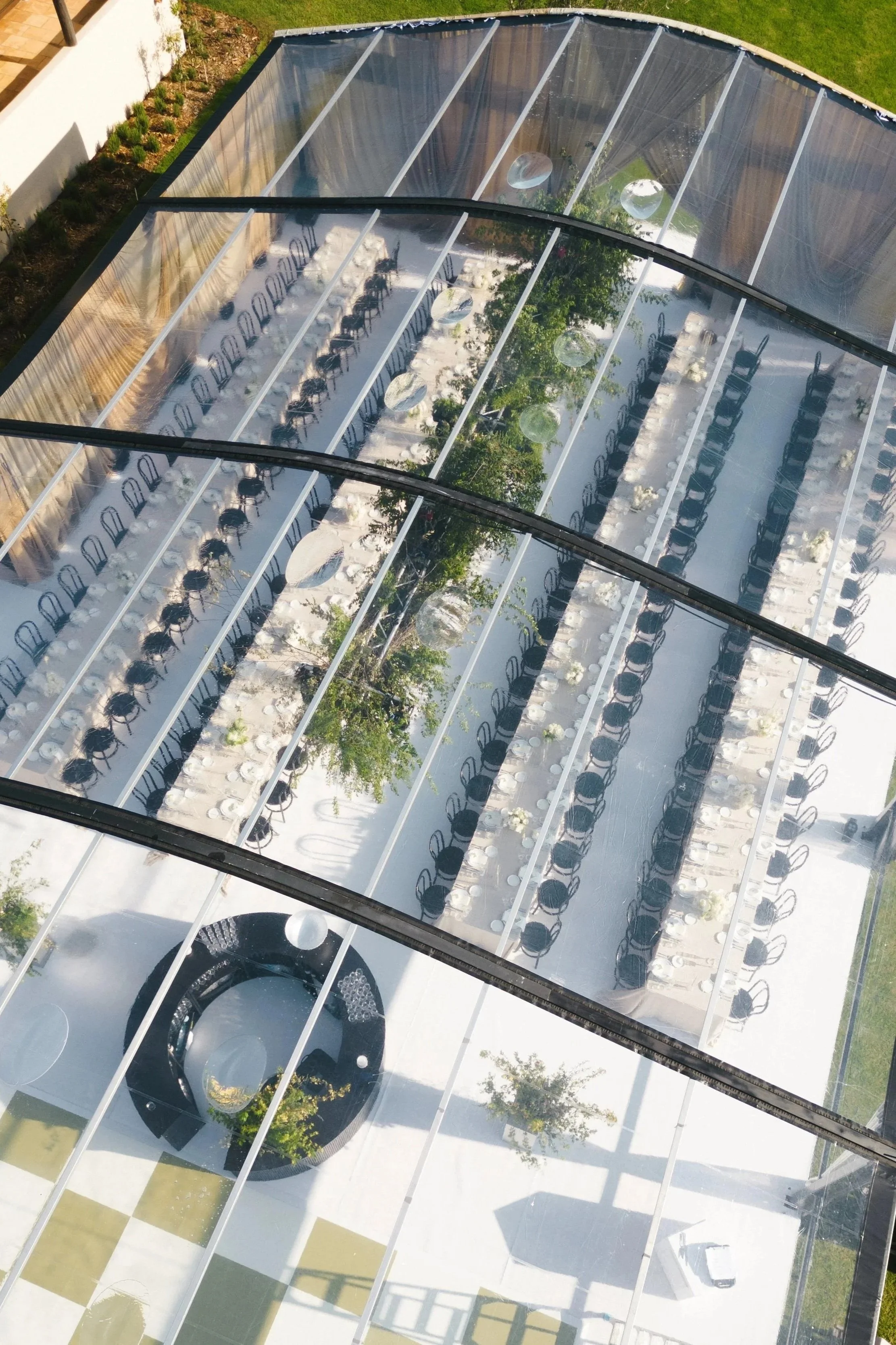 Aerial view of a modern building with a glass roof, outdoor dining area with tables and chairs, and a garden with trees and grass.