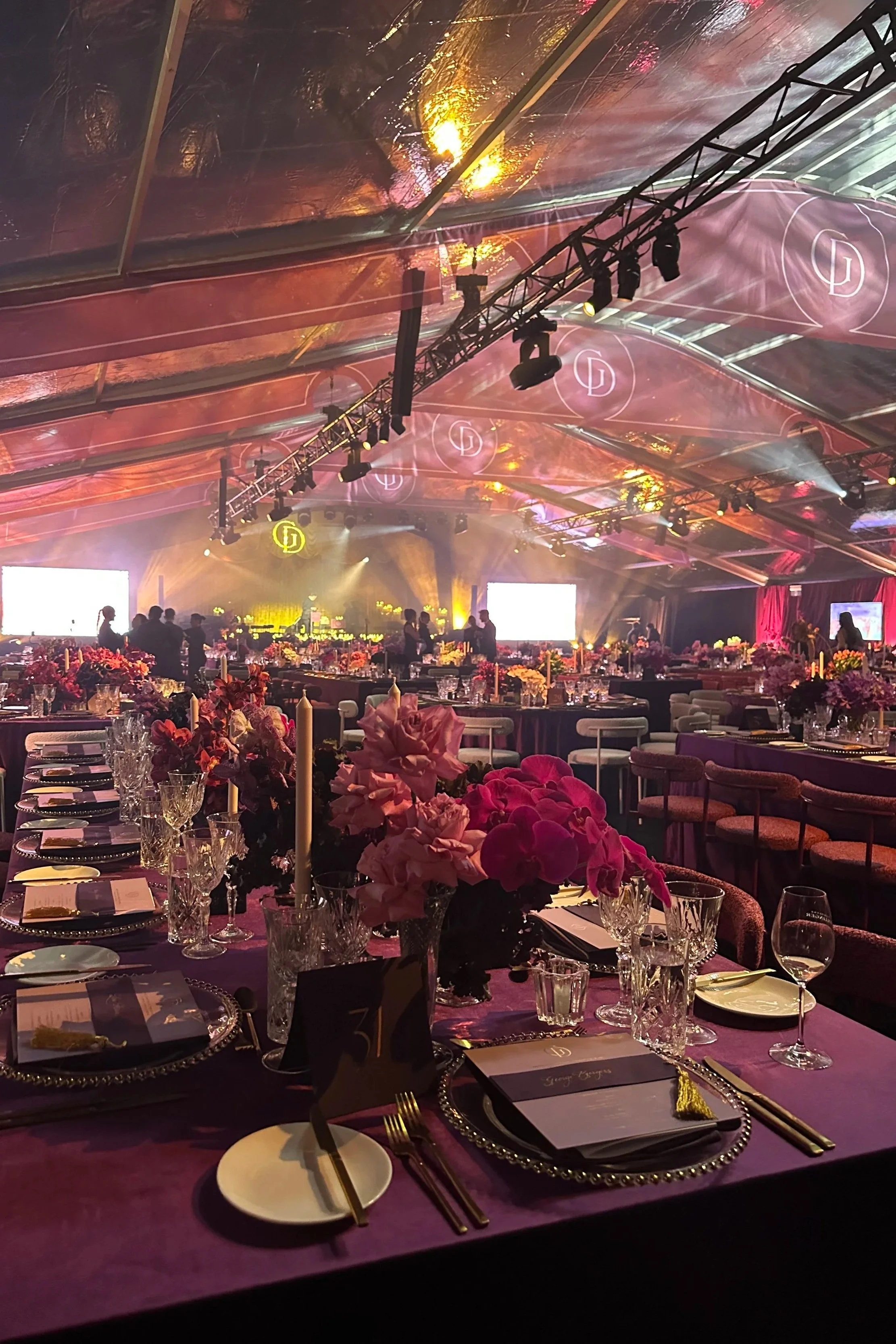 Elegant event space with purple tablecloths, floral centerpieces, ornate plates, glassware, and cutlery. The ceiling features hanging lights, decorative branding logos, and professional lighting equipment. Guests and staff are visible in the backgrou