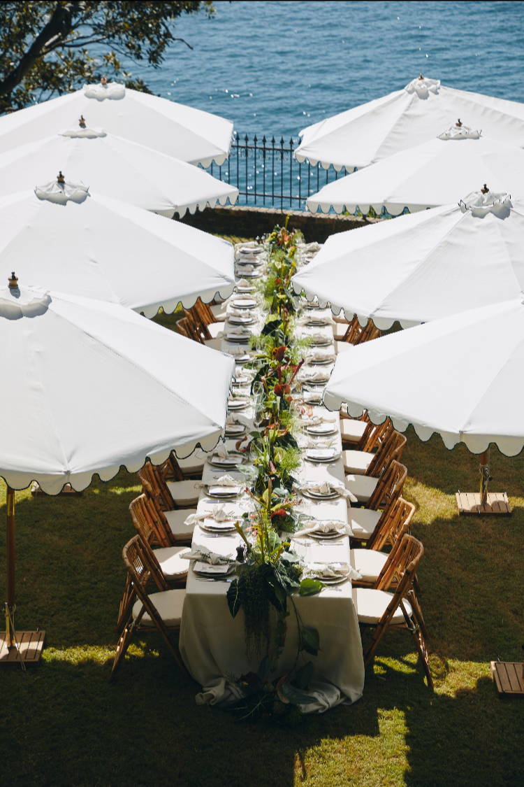 Long outdoor dining table set for a formal event with white tablecloth, plates, glasses, and floral centerpieces, surrounded by wooden chairs, shaded by large white umbrellas, overlooking a body of water.