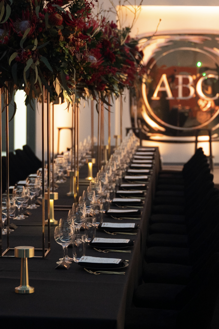 Elegant event setup with a long black table, tall gold candleholders, floral centerpieces, and place settings including wine glasses and printed menus, in a warmly lit room with a large illuminated sign reading 'ABC' in the background.