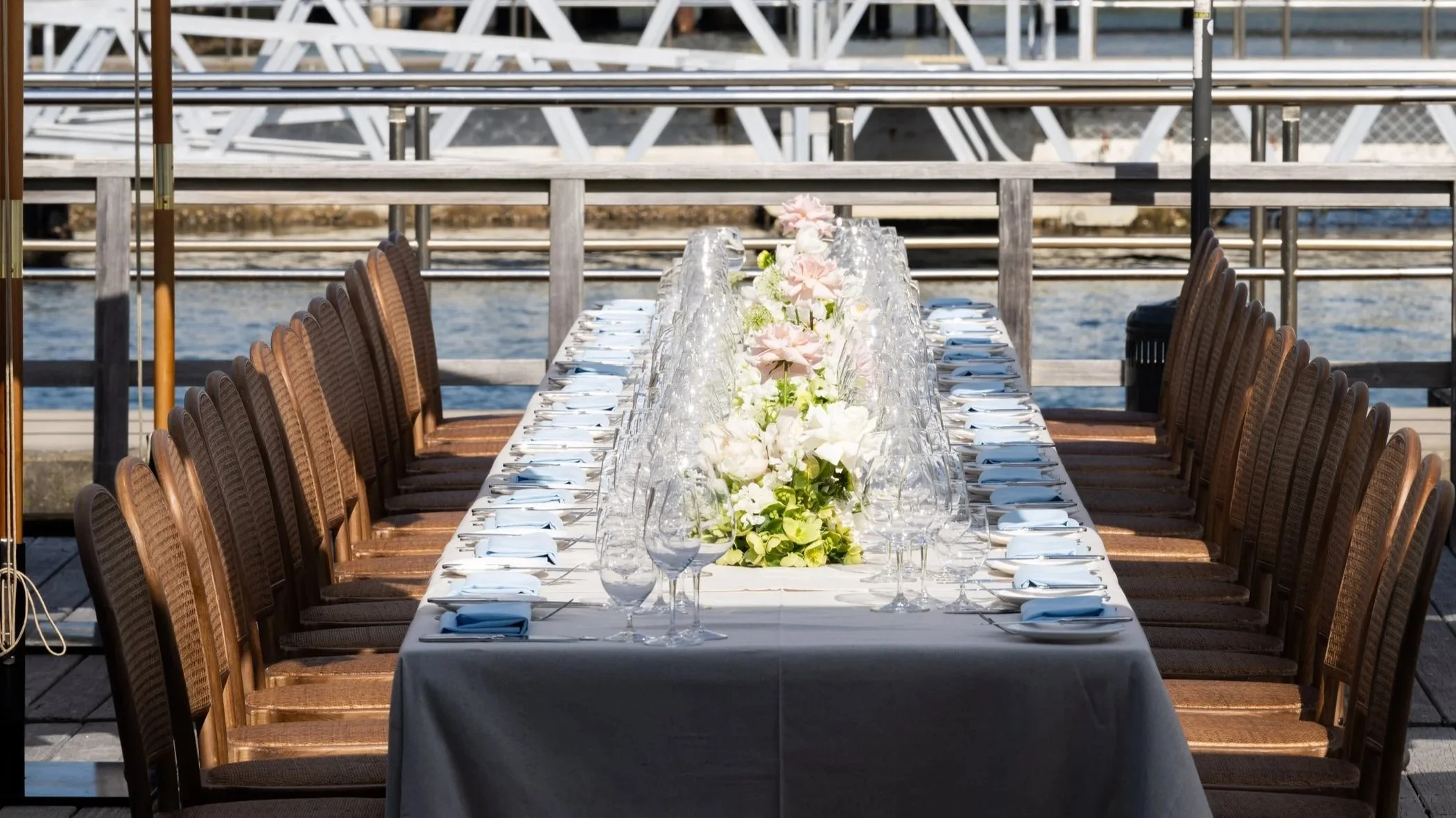 A long outdoor dining table set with white tablecloths, glassware, plates, and floral centerpieces, located on a wooden deck by the water with docks in the background.