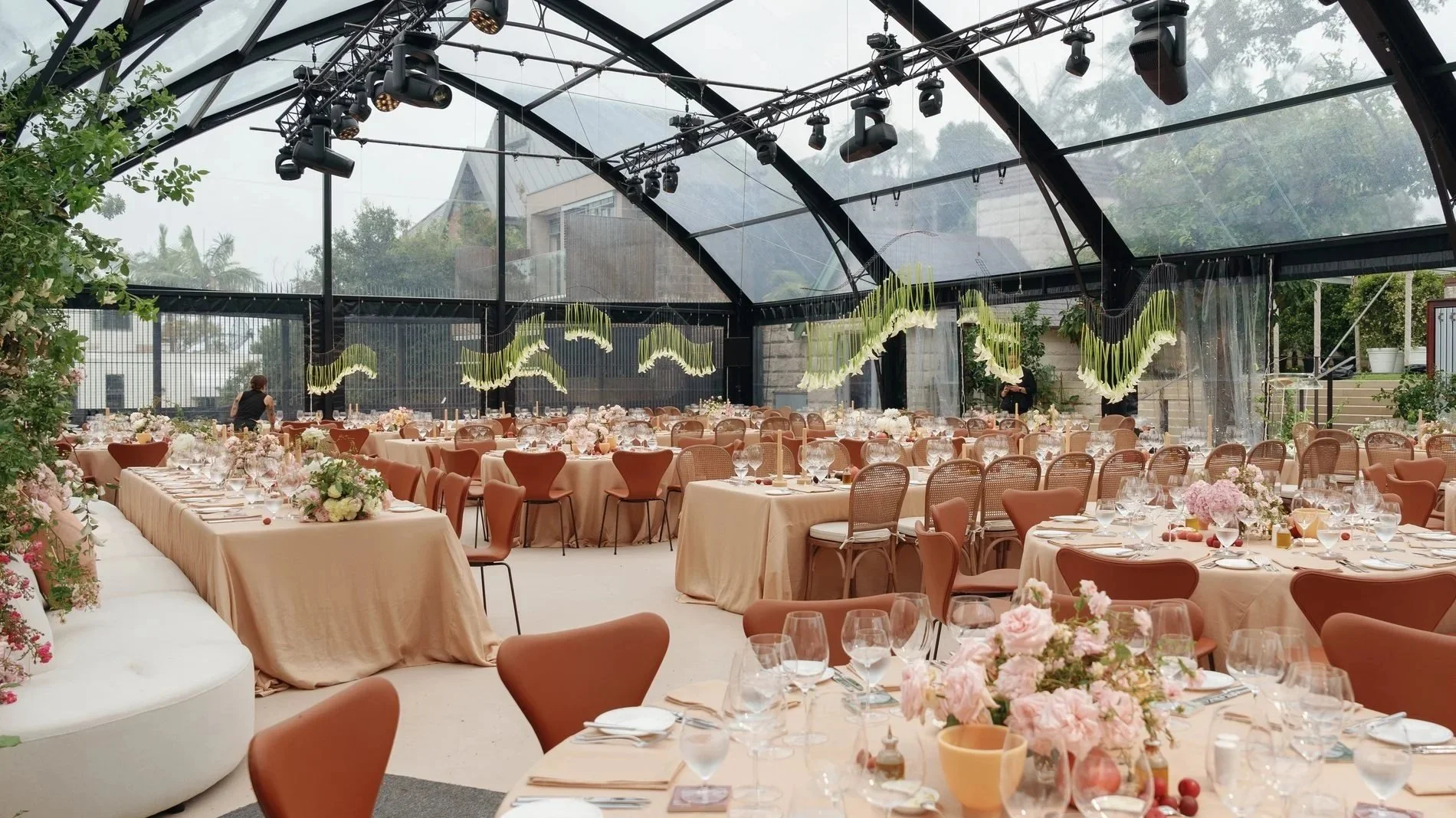 Elegant indoor wedding reception with round and long banquet tables covered in beige tablecloths, decorated with pink and white floral centerpieces, and set with glassware, plates, and cutlery. The space features a transparent ceiling, black metal fr