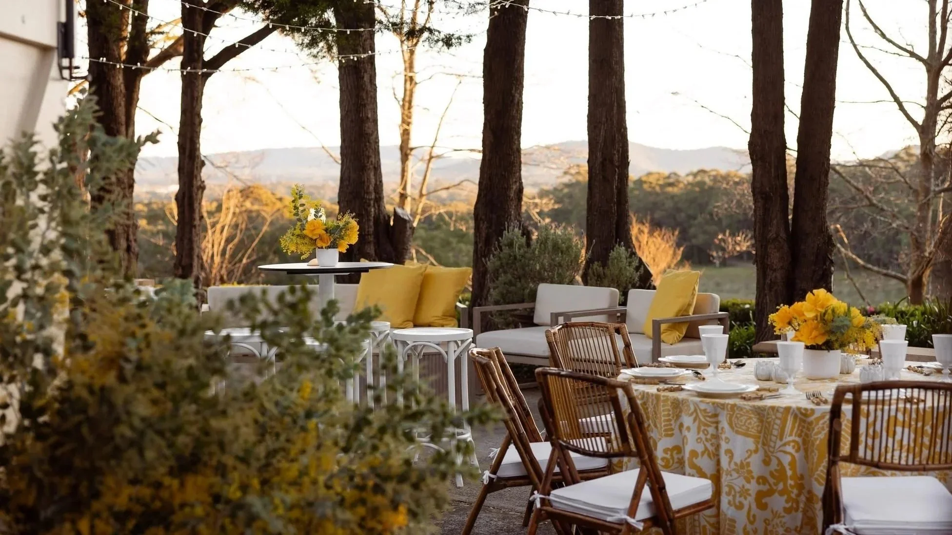 An outdoor dining area with a table covered with a yellow patterned tablecloth, surrounded by various chairs, and decorated with yellow flowers in vases. The setting is among trees with fairy lights overhead, overlooking a scenic landscape at sunset.
