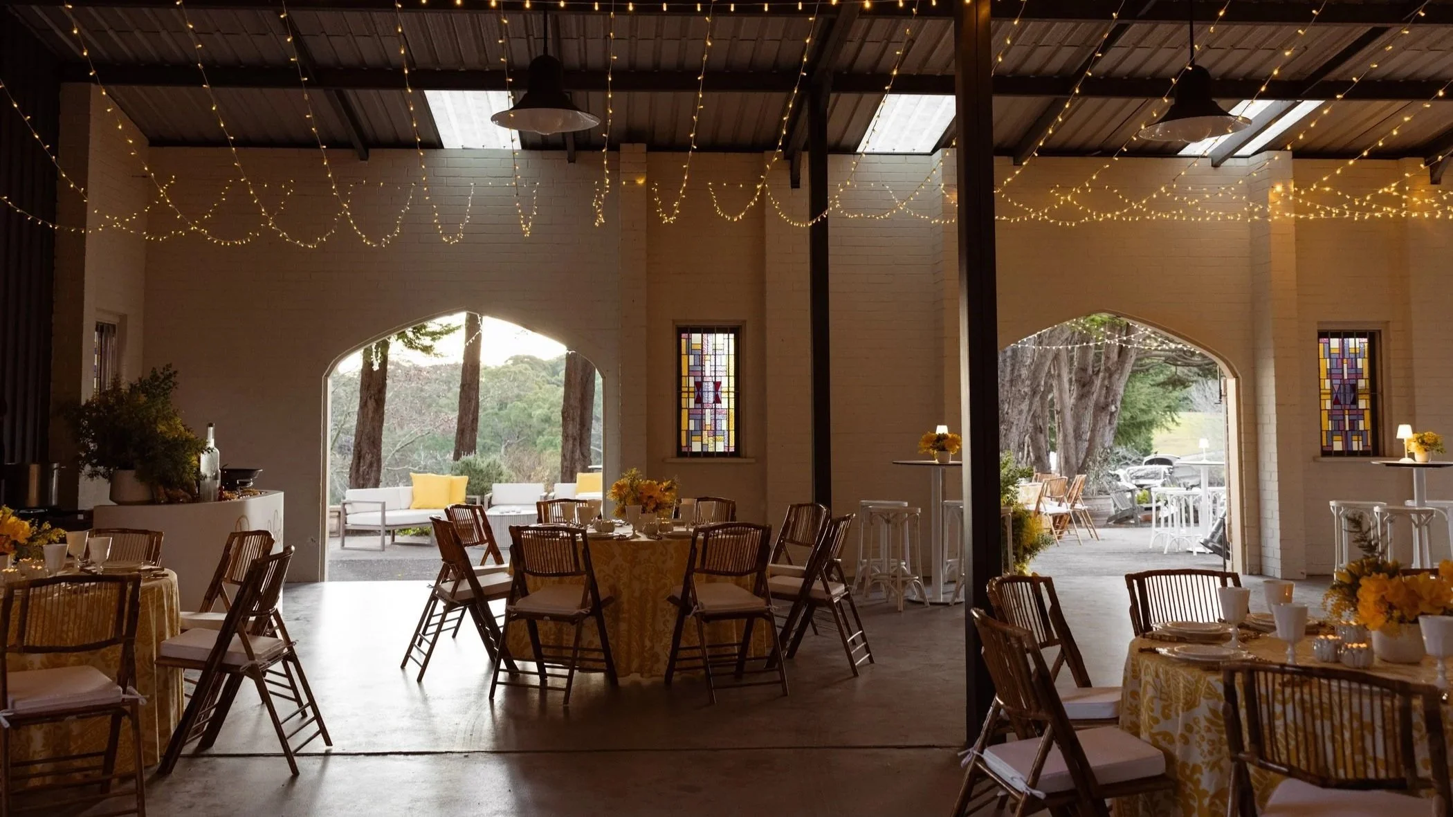 Interior of a rustic event space decorated with string lights hanging from the ceiling, tables with yellow floral centerpieces, and chairs arranged for a celebration. Large open doors reveal an outdoor patio with trees and seating.
