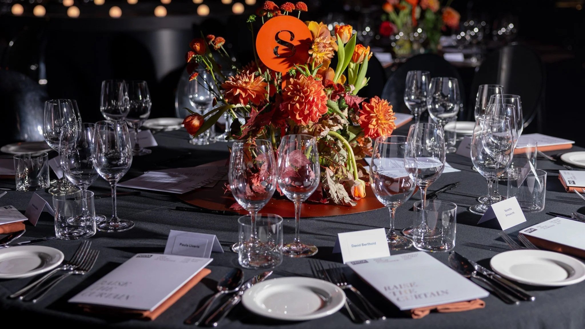 Table set for a formal event with a floral centerpiece in shades of orange, red, and yellow, surrounded by wine glasses, plates, and utensils on a dark tablecloth.