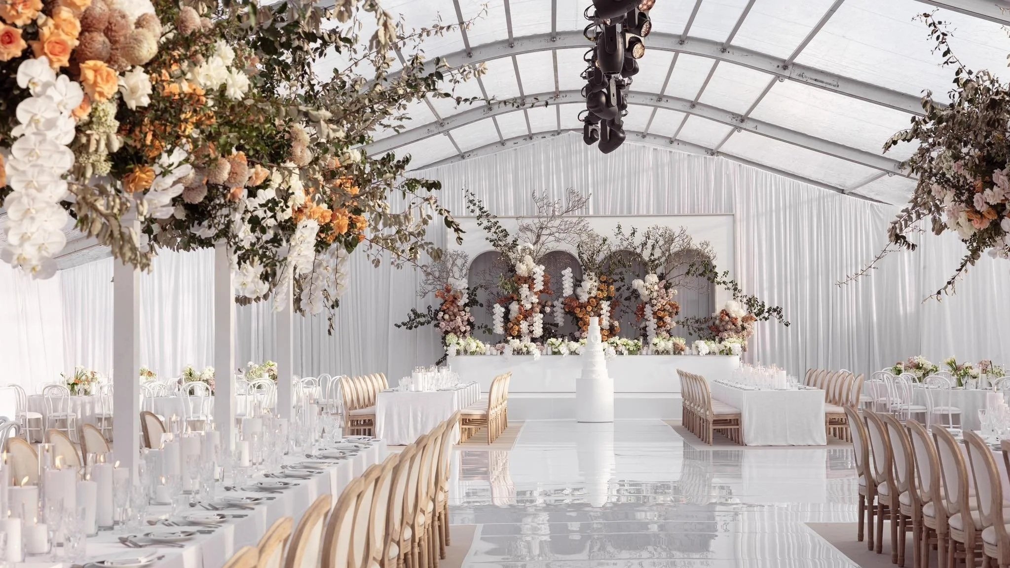 Elegant wedding reception setup inside a white canopy tent with floral decorations and long tables with white tablecloths.