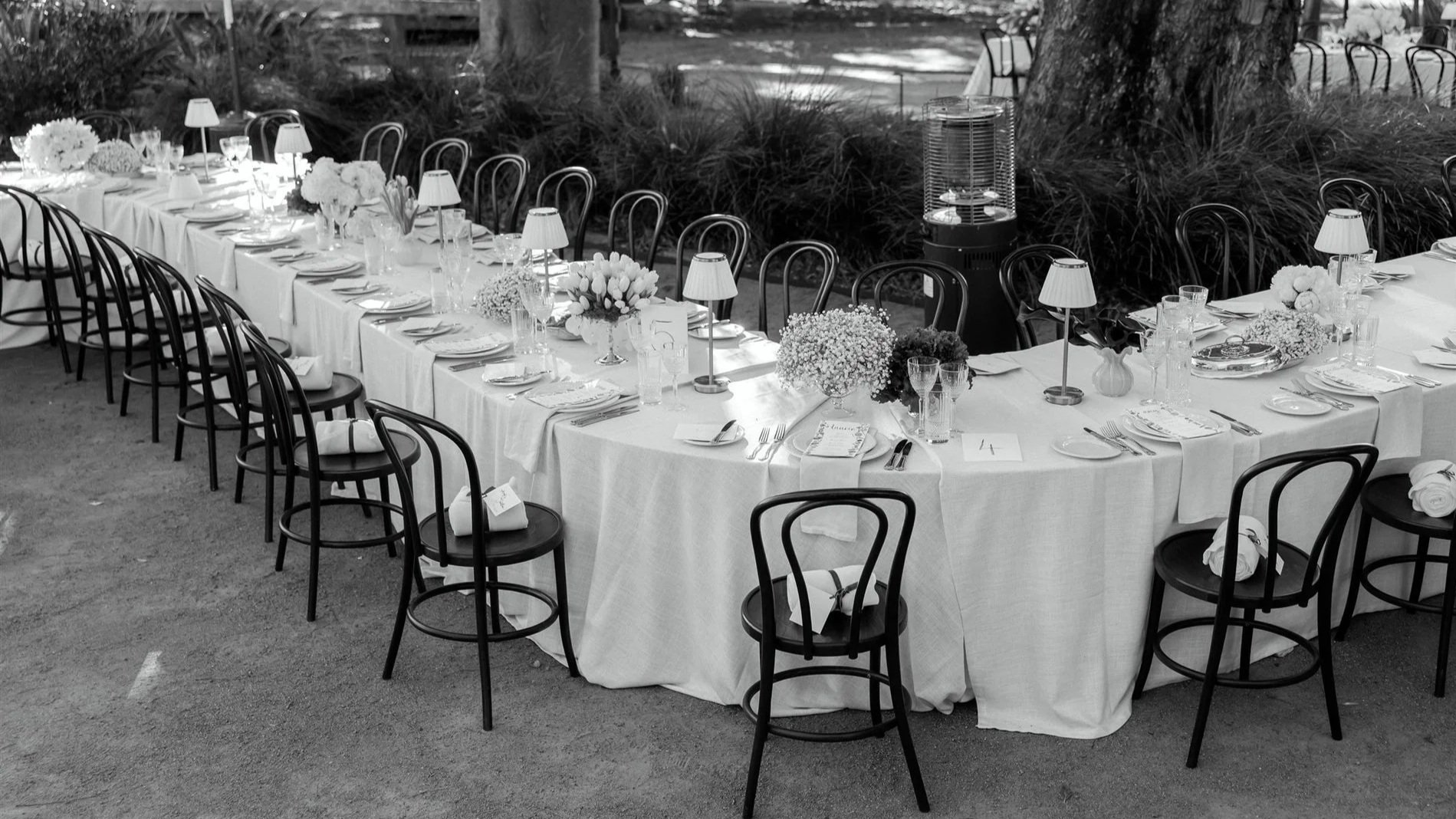 Long banquet table set for a formal event with white tablecloths, floral centerpieces, small table lamps, plates, silverware, glasses, and napkins. Black chairs surround the table, with greenery and trees in the background.