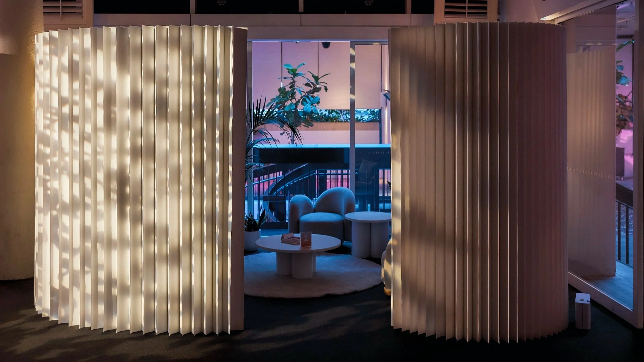Interior view of a modern lounge or waiting area with illuminated slatted partition walls, a small round table, an armchair, potted plants, and a balcony with glass doors and railings, illuminated by colorful ambient lighting.